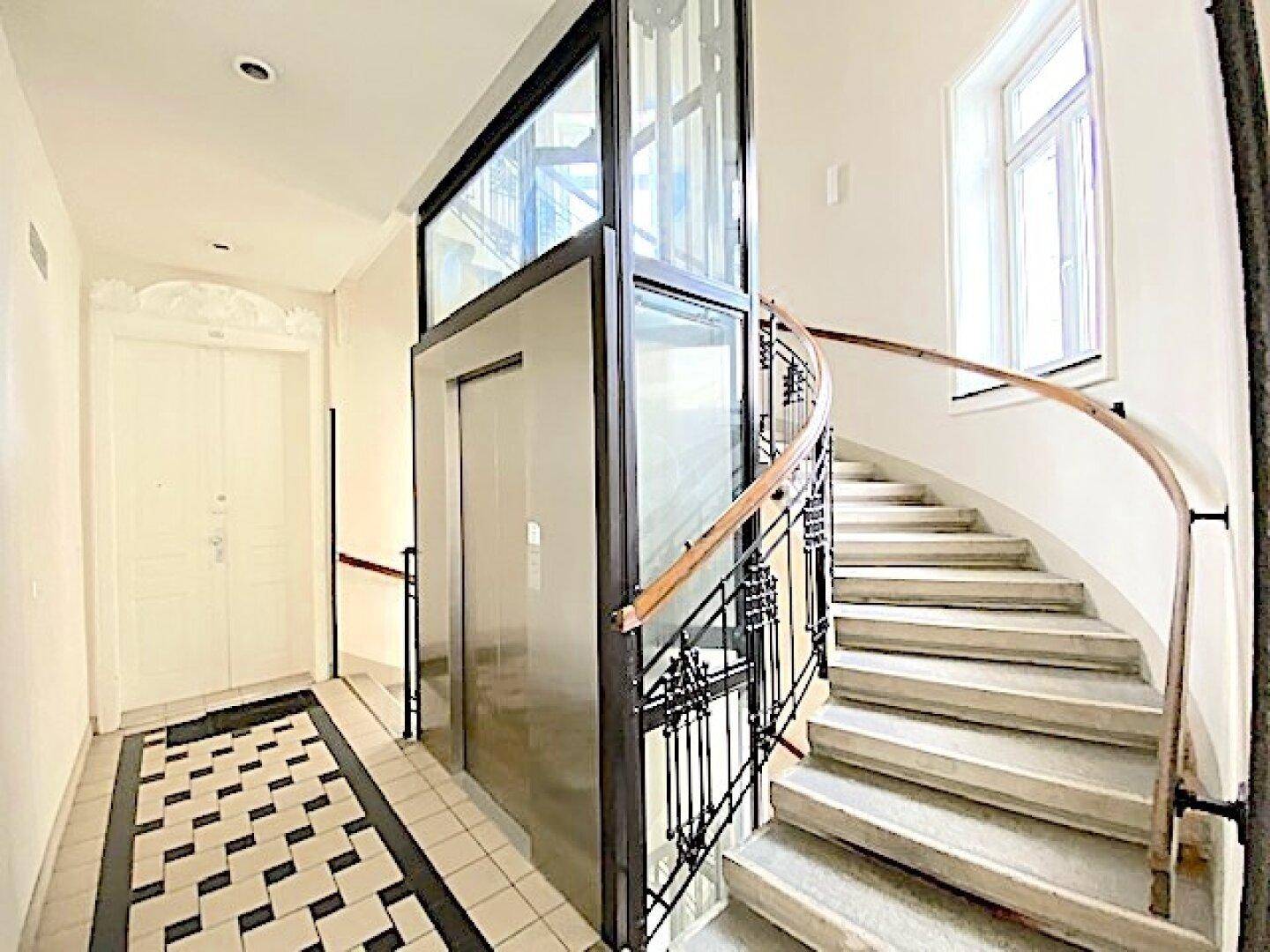Heller Eingangsbereich mit modernem Glasaufzug, geschwungener Treppe und gemustertem Fliesenboden.