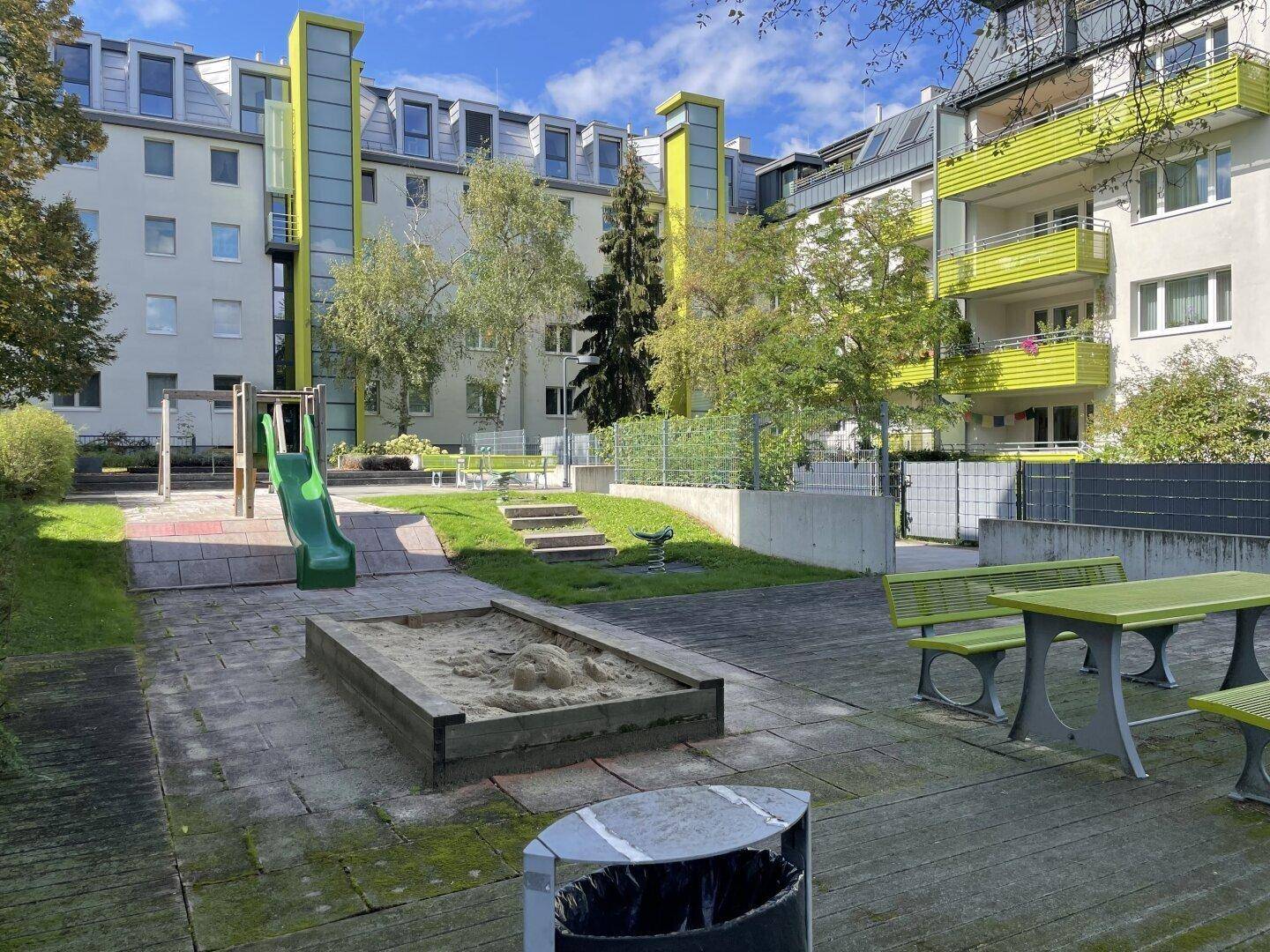 Gemeinschaftlicher Spielplatz mit Sandkasten und Rutsche, umgeben von Grünflächen und Wohngebäuden.