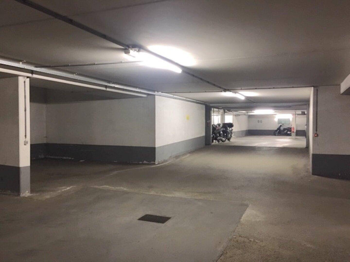 Weitläufige Tiefgarage mit mehreren Stellplätzen und Motorrädern im Hintergrund, gut beleuchtet.