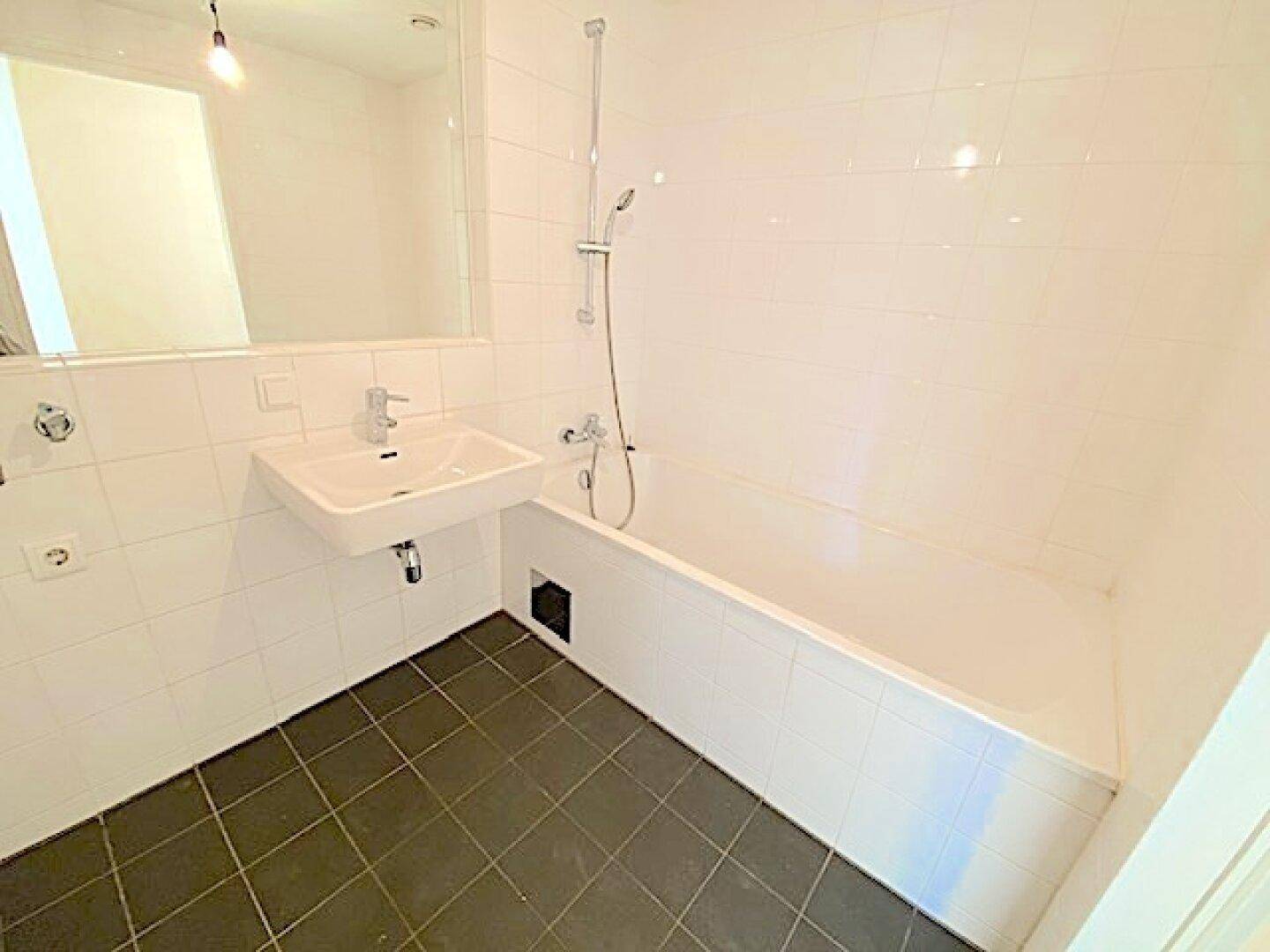 Badezimmer mit Badewanne, Waschbecken und weißen Wandfliesen, ergänzt durch dunkle Bodenfliesen.