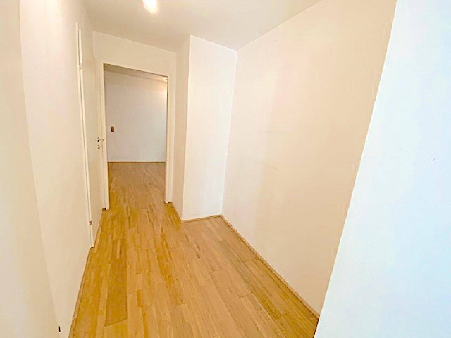 Schmaler, heller Flur mit Holzboden und weißen Wänden, der einen Durchgang zu weiteren Zimmern bietet.