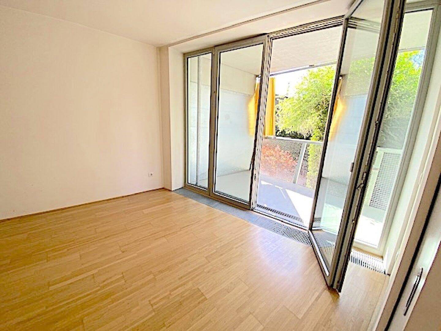 Heller Raum mit Holzboden und einer breiten Glasfront, die Zugang zu einem Außenbereich bietet.