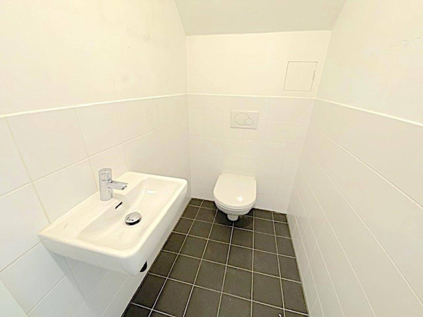 Modernes Gäste-WC mit wandhängendem Waschbecken und Toilette, weißen Fliesen und dunklem Boden.