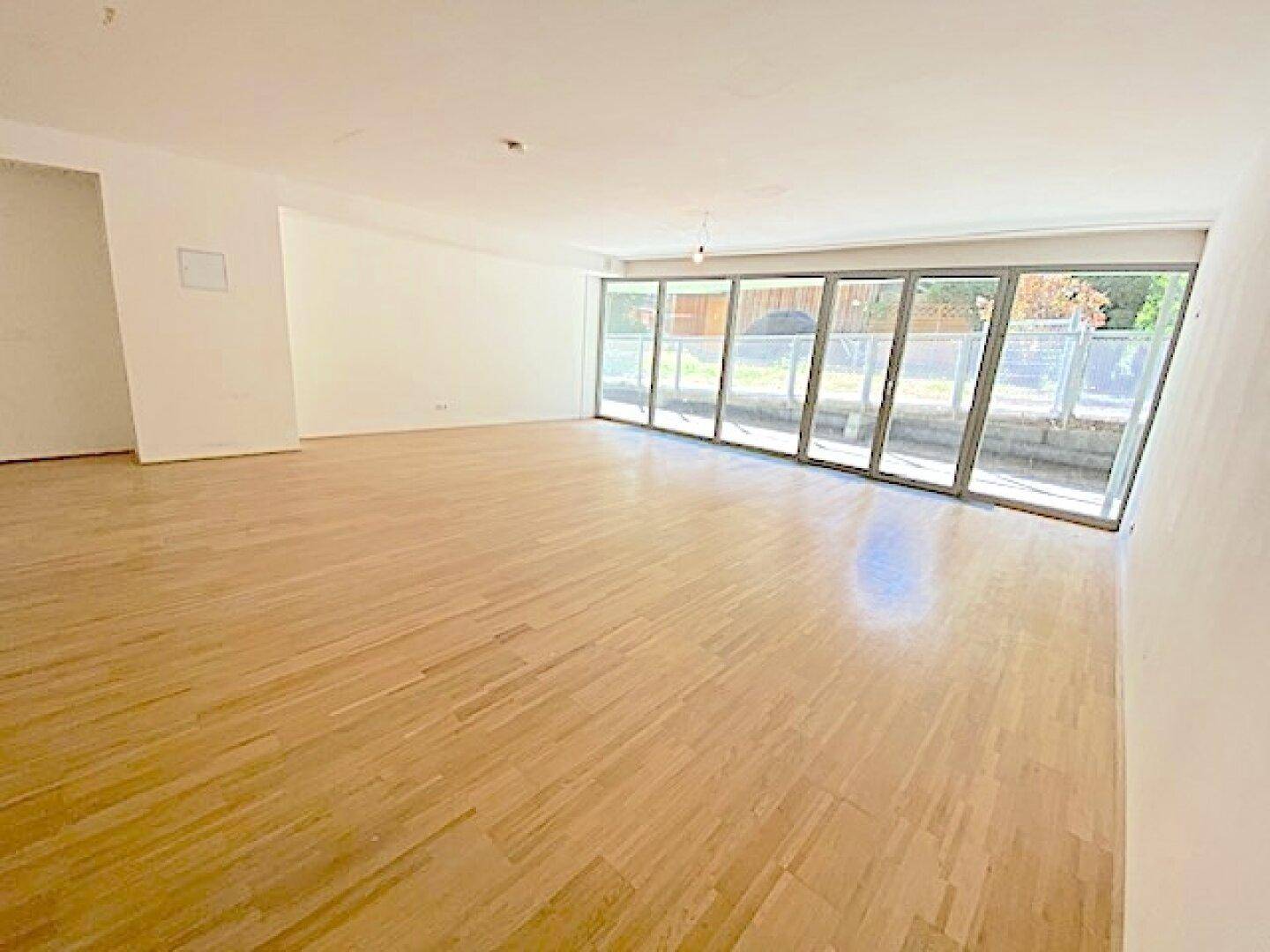 Geräumiger Wohnbereich mit Holzboden und einer langen Fensterfront, die den Raum hell erscheinen lässt.