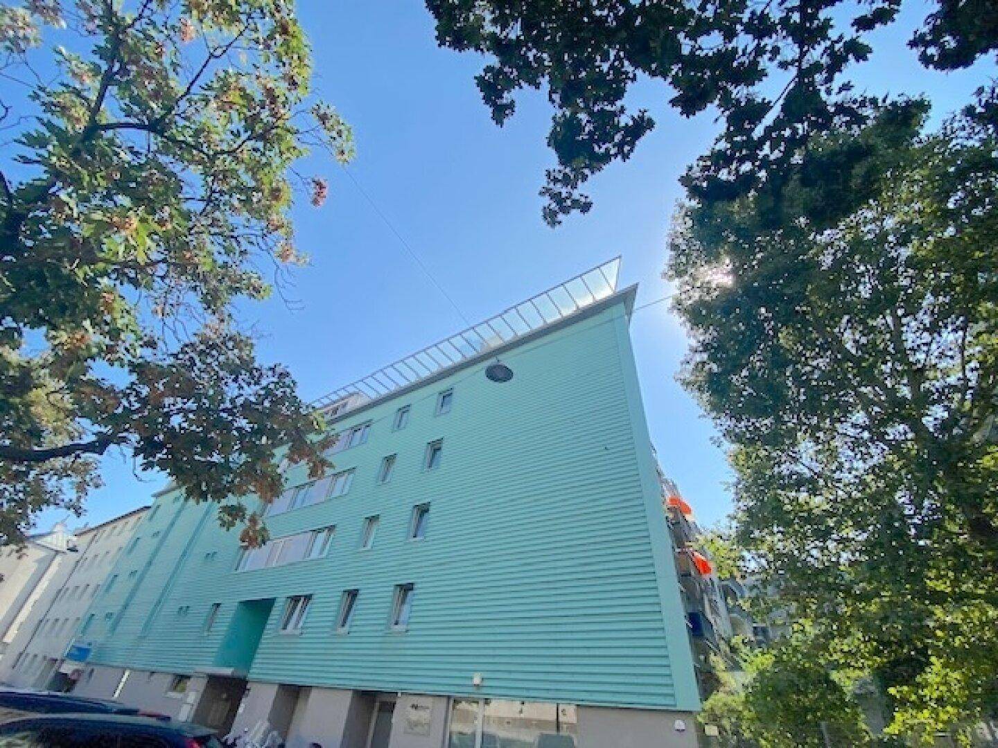 Moderne Gebäudefassade in Türkis mit vielen Fenstern und umgebenden Bäumen unter blauem Himmel.