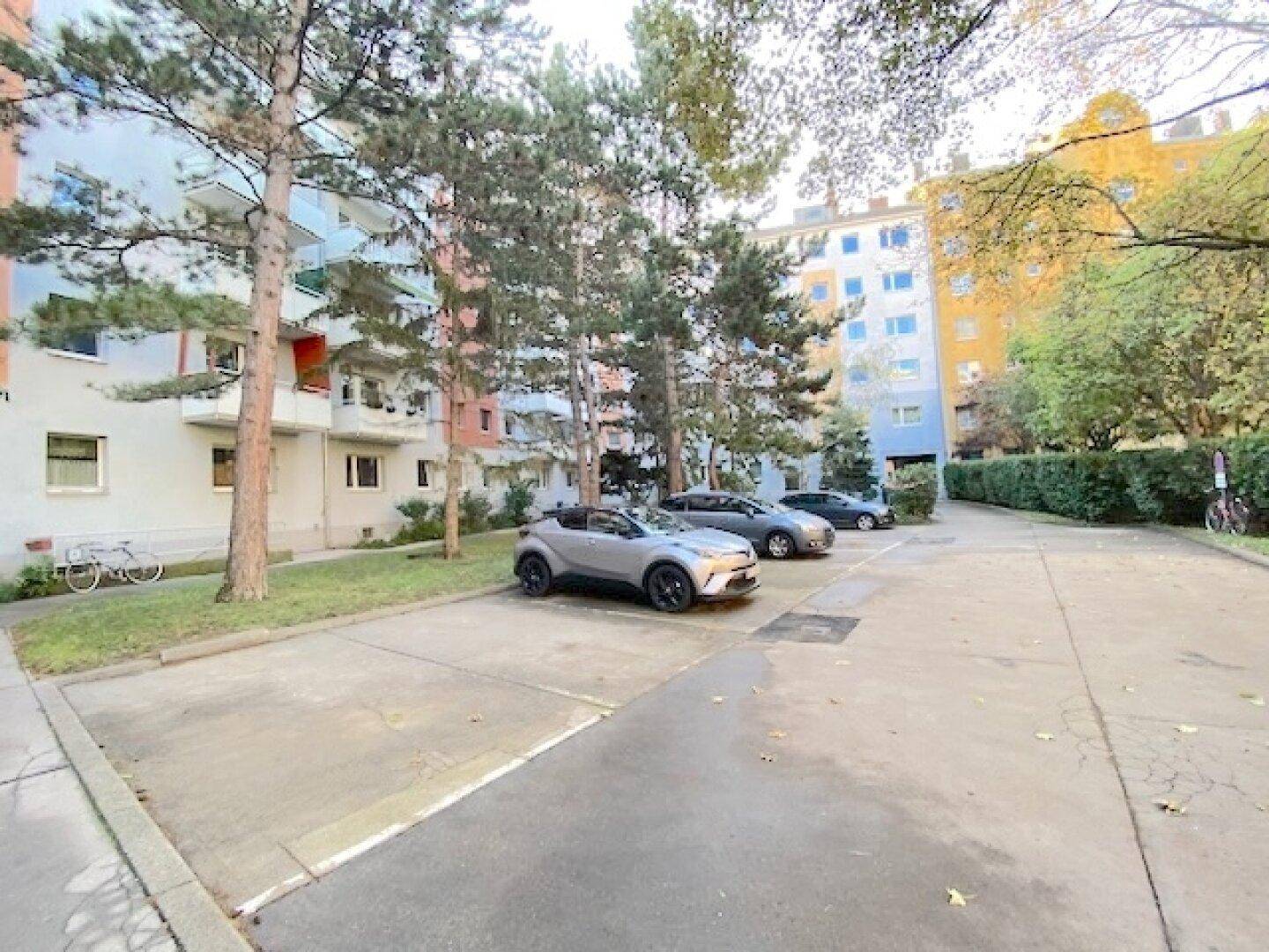 Außenparkplätze vor einem Wohngebäude, umgeben von Bäumen und Grünflächen.