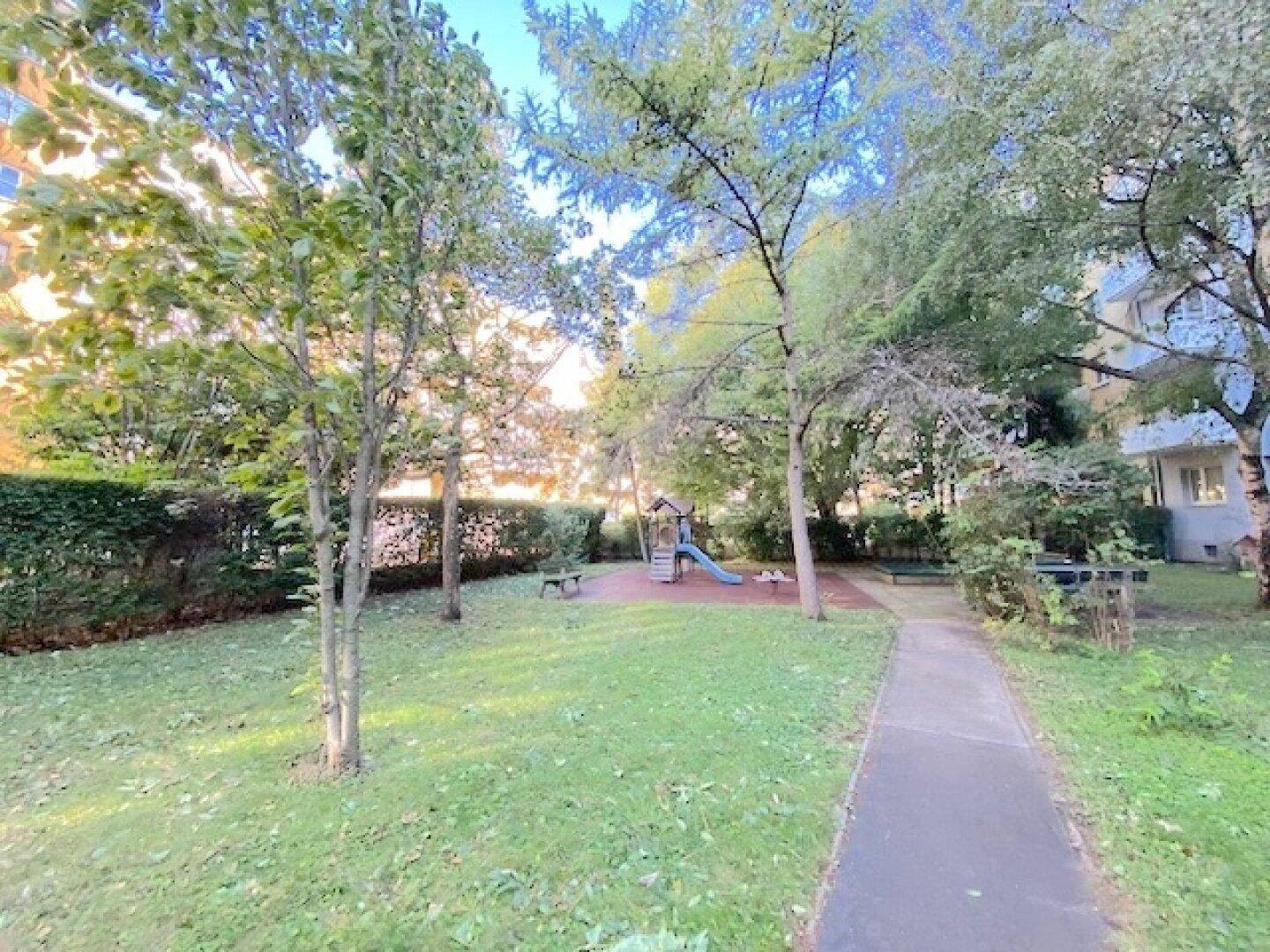 Grüner Gemeinschaftsgarten mit Spielplatz und Rutsche, umgeben von Bäumen.