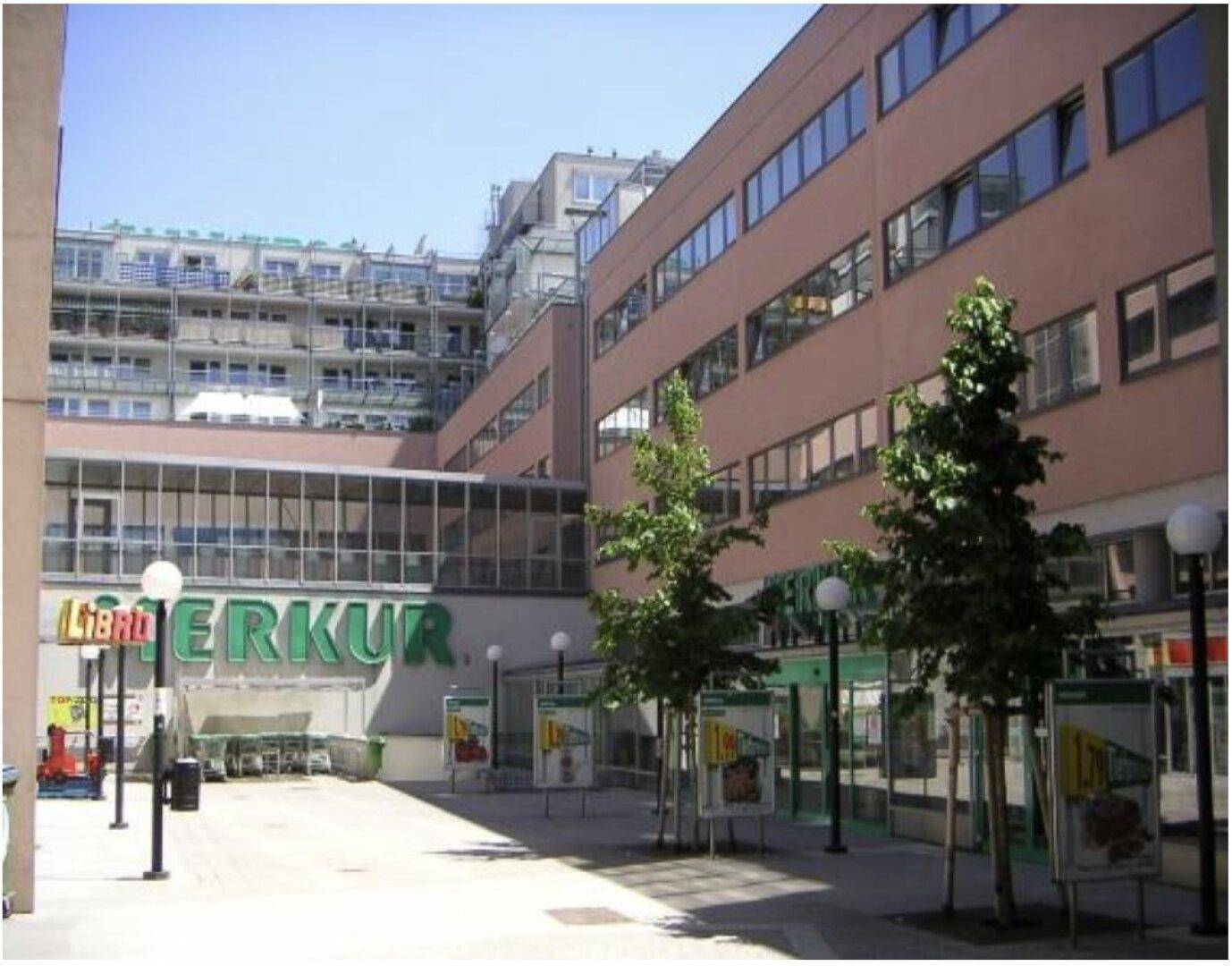 Gebäudekomplex mit Geschäftsflächen und Wohnungen, umgeben von Bäumen und Laternen.