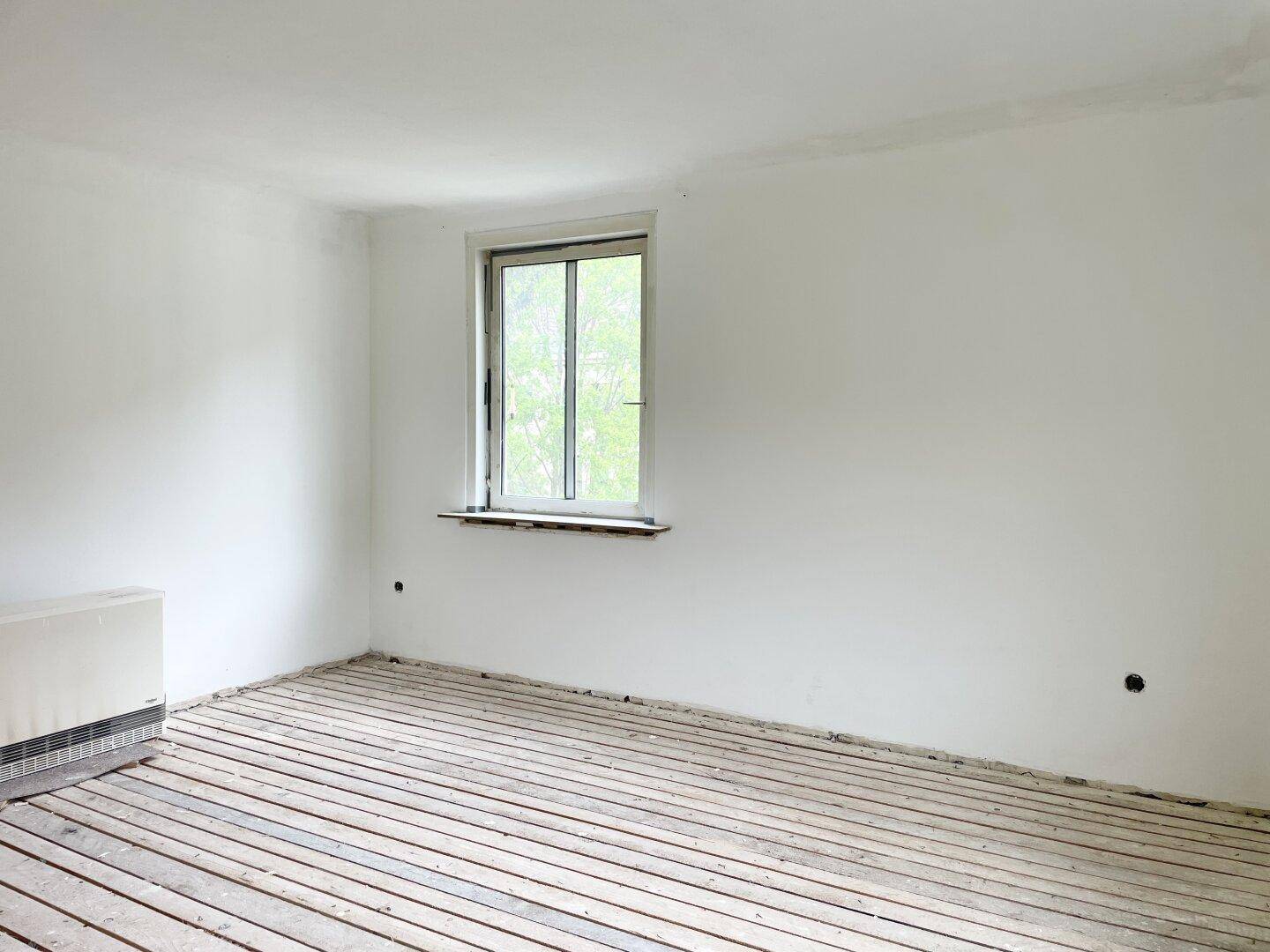 Renovierungsbedürftiger Raum mit Heizkörper, weiß gestrichenen Wänden und Blick ins Grüne.