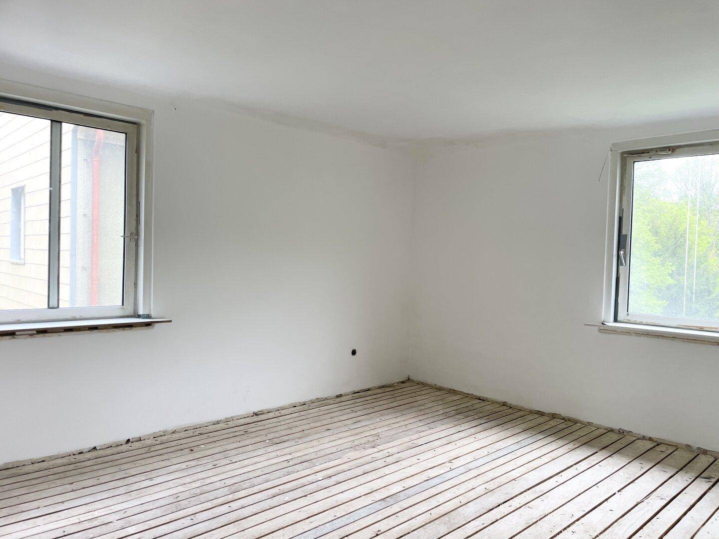 Heller Raum mit zwei Fenstern, weiß gestrichenen Wänden und freigelegten Holzdielen.