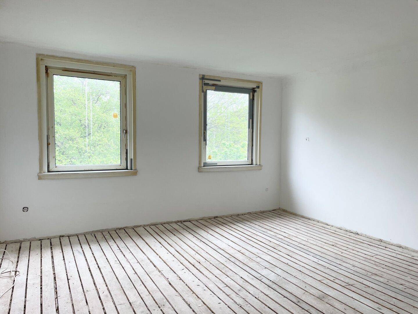 Leerer Raum mit zwei Fenstern, die einen Blick ins Grüne bieten, und Holzdielenboden.