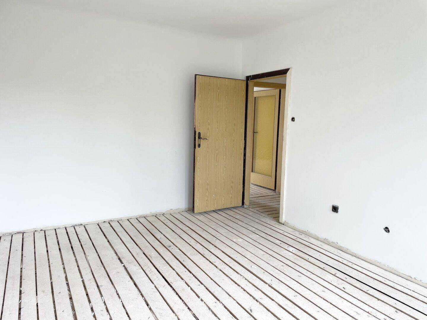 Leerer Raum mit weiß gestrichenen Wänden und offener Holztür, die zu einem weiteren Zimmer führt.