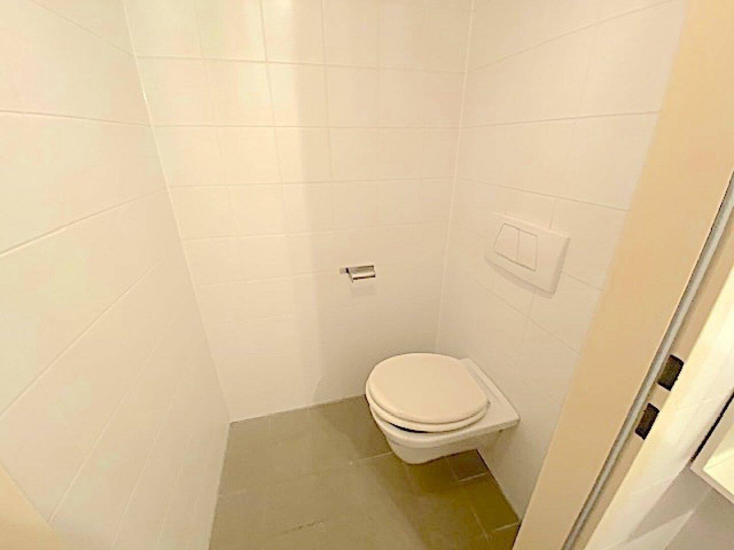 Kleiner, sauberer Toilettenraum mit weißen Wandfliesen und einer wandhängenden Toilette.