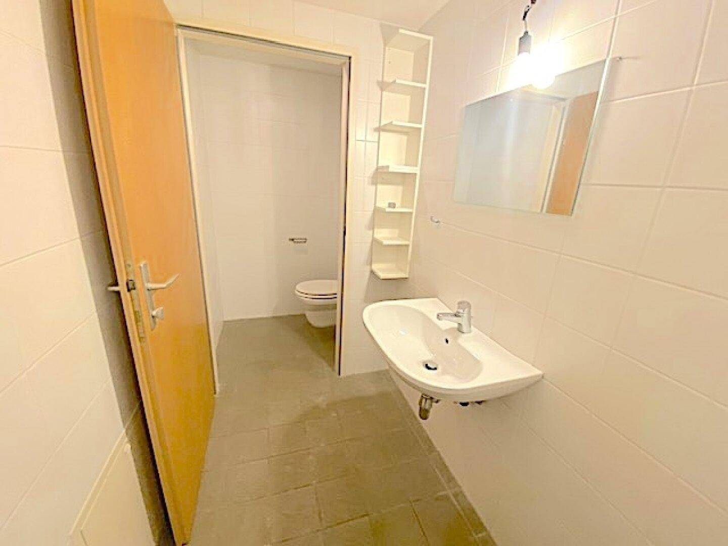 Badezimmer mit Waschbecken, Spiegel und Zugang zu einer separaten Toilette, alles hell gefliest.