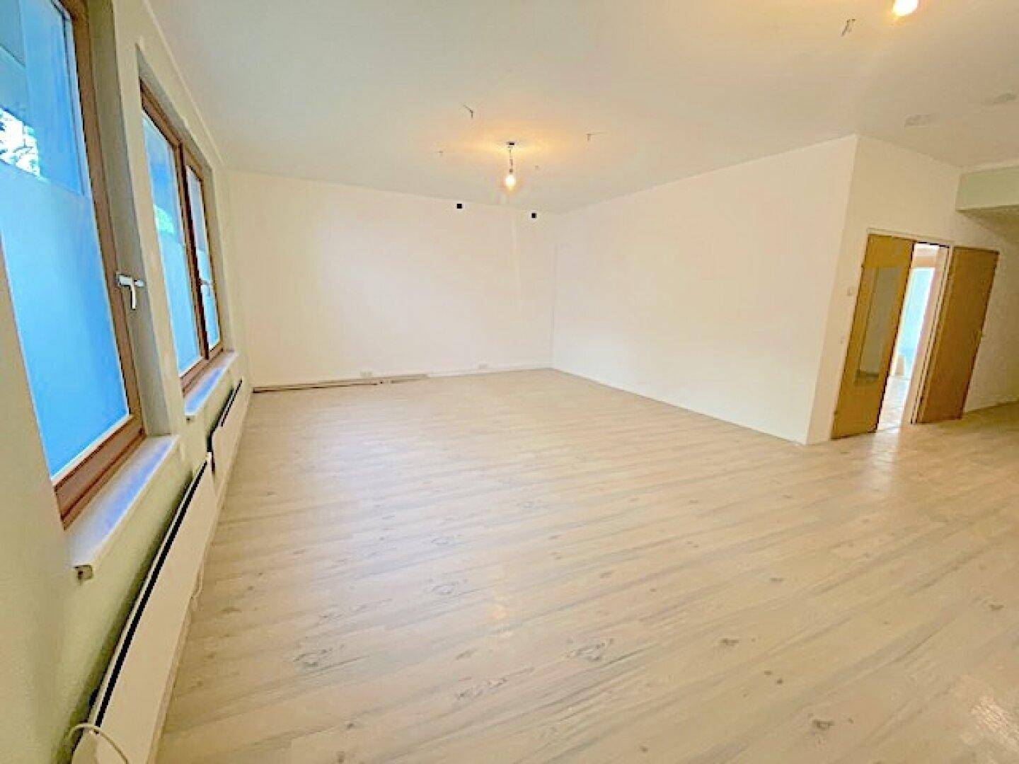 Geräumiger Raum mit hellem Laminatboden und großen Fenstern, die viel Licht hereinlassen.