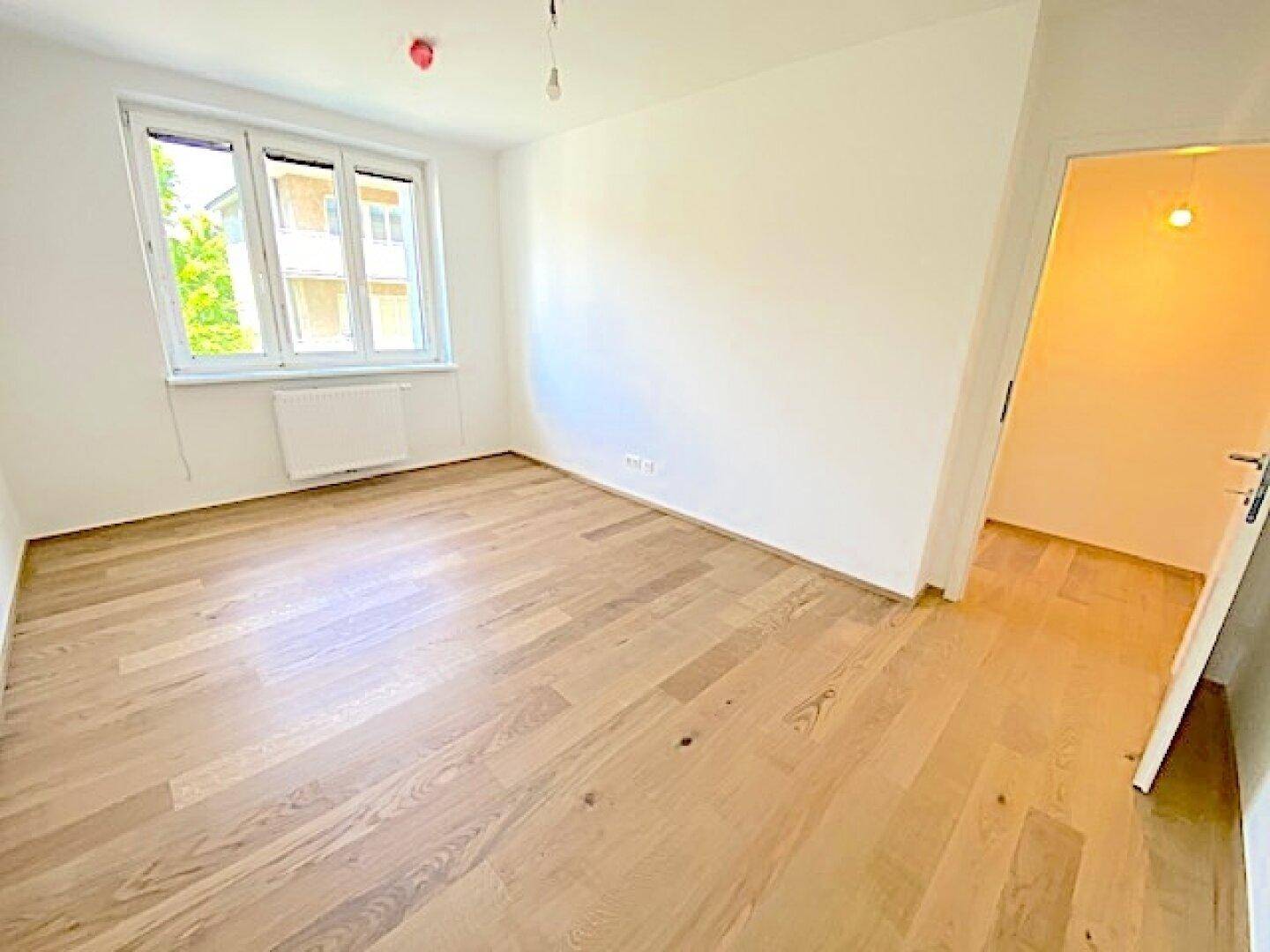 Geräumiges, leeres Zimmer mit Holzboden und Fenster, bietet viel Tageslicht.
