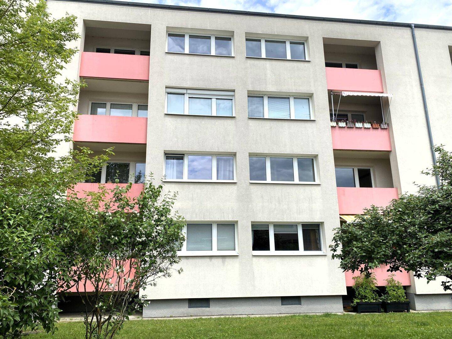 Mehrfamilienhaus mit heller Fassade und rosa Balkonen, umgeben von gepflegten Grünflächen und Bäumen.