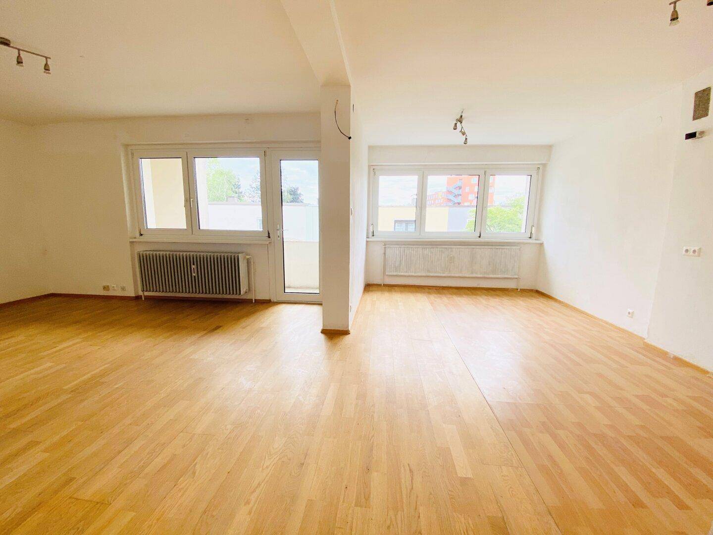 Geräumiger Wohnbereich mit Parkettboden und großen Fenstern, die viel Licht hereinlassen.