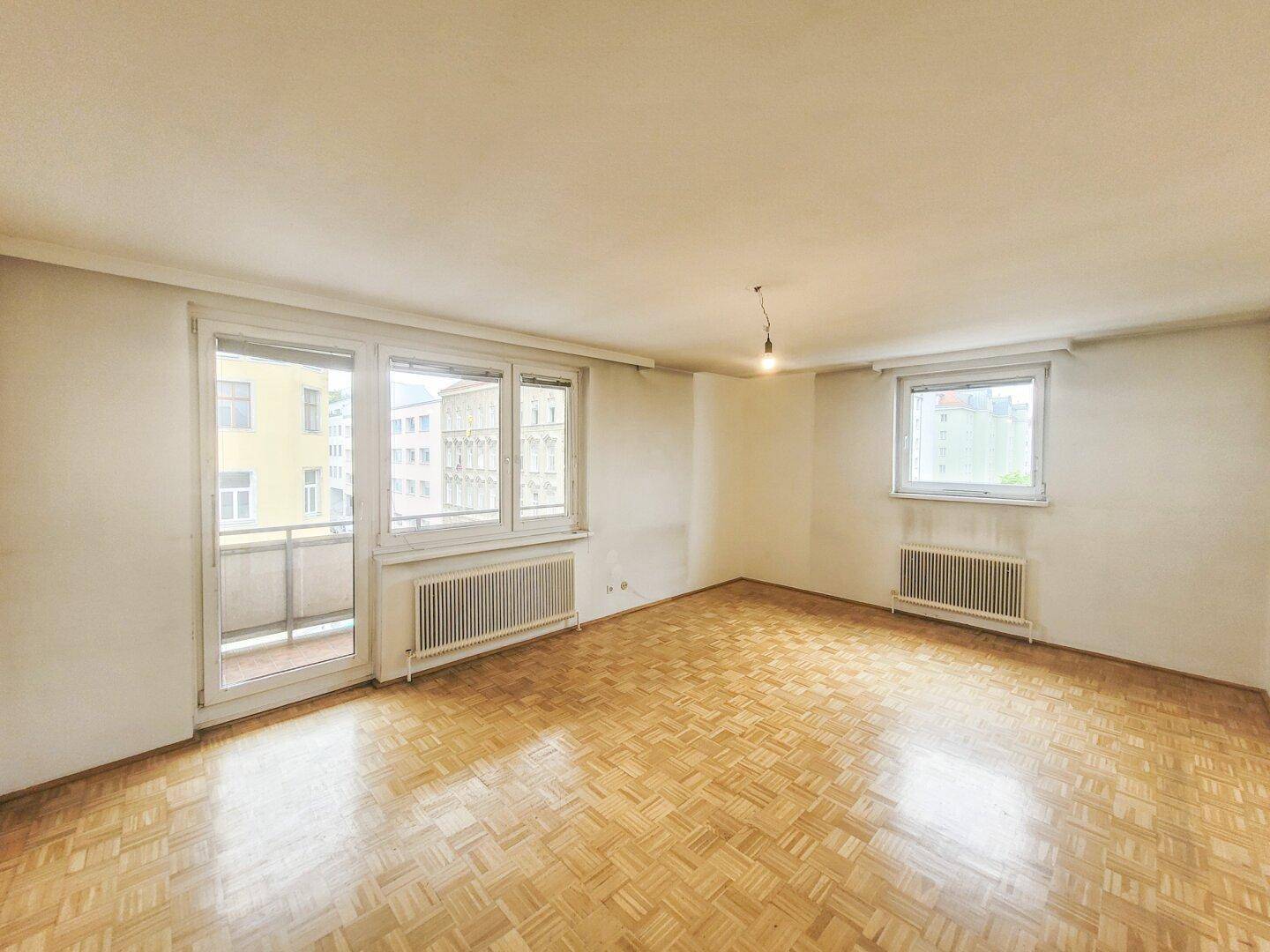 Helles Zimmer mit Parkettboden, weißen Wänden und Zugang zu einem Balkon.