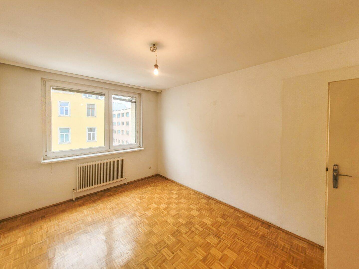 Helles Schlafzimmer mit Parkettboden, weißen Wänden und einem Fenster zur Straße.