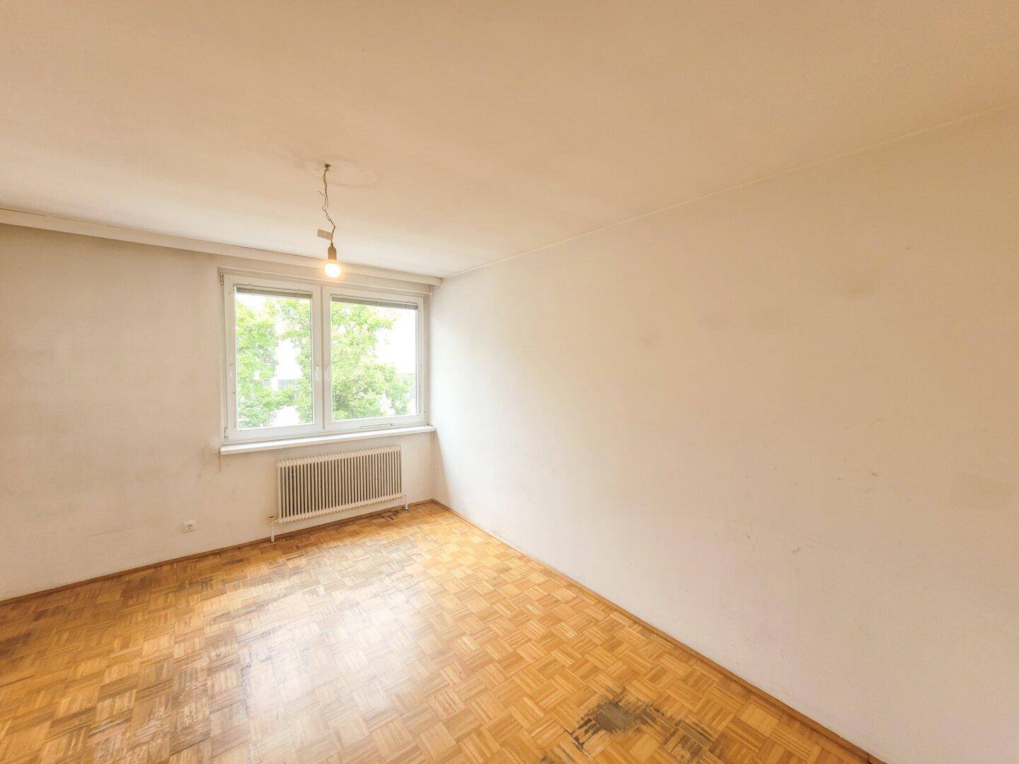Ruhiges Zimmer mit Parkettboden, weißen Wänden und Blick ins Grüne.