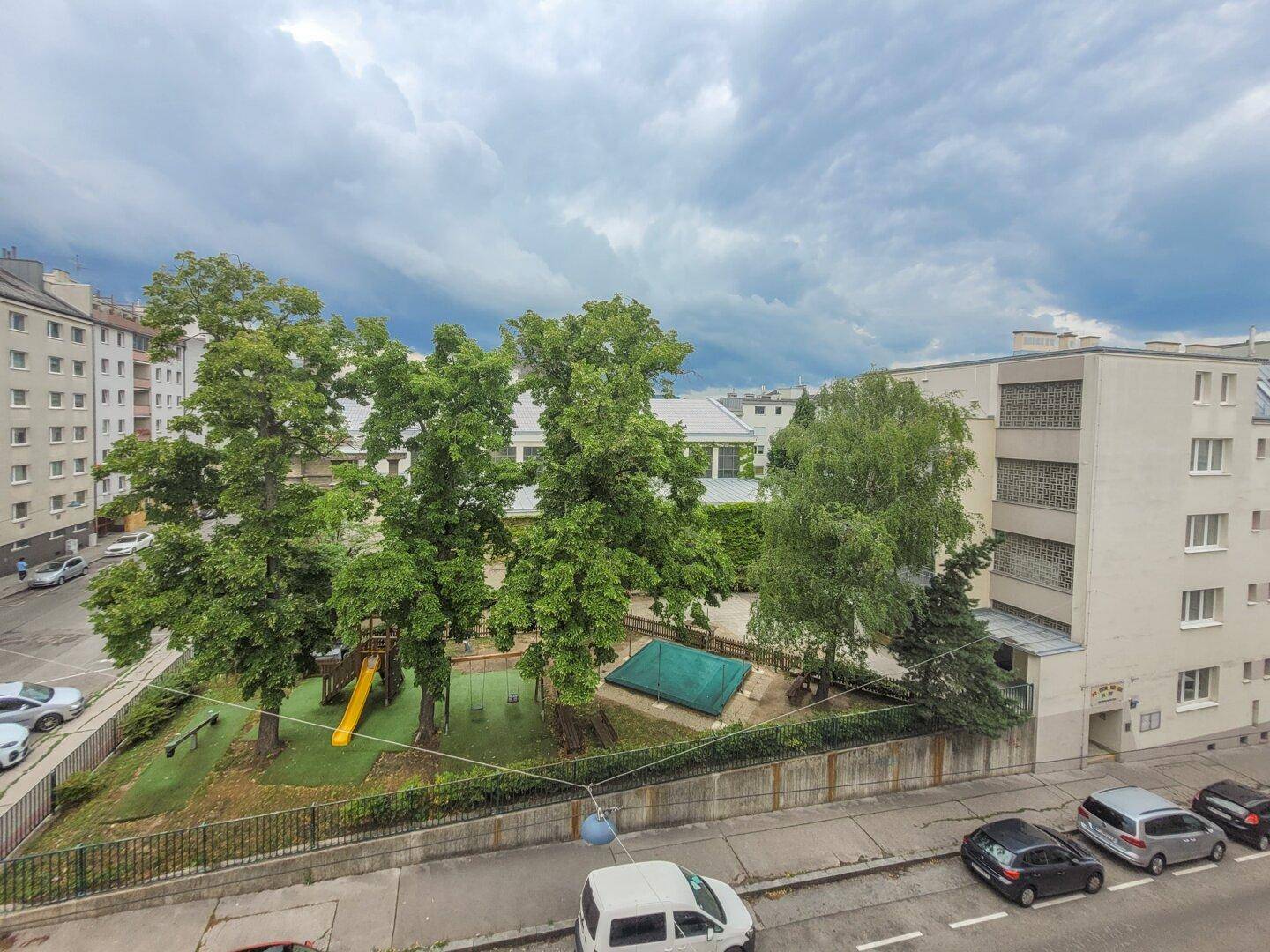 Blick auf einen grünen Innenhof mit Spielplatz und hohen Bäumen, umgeben von Gebäuden.