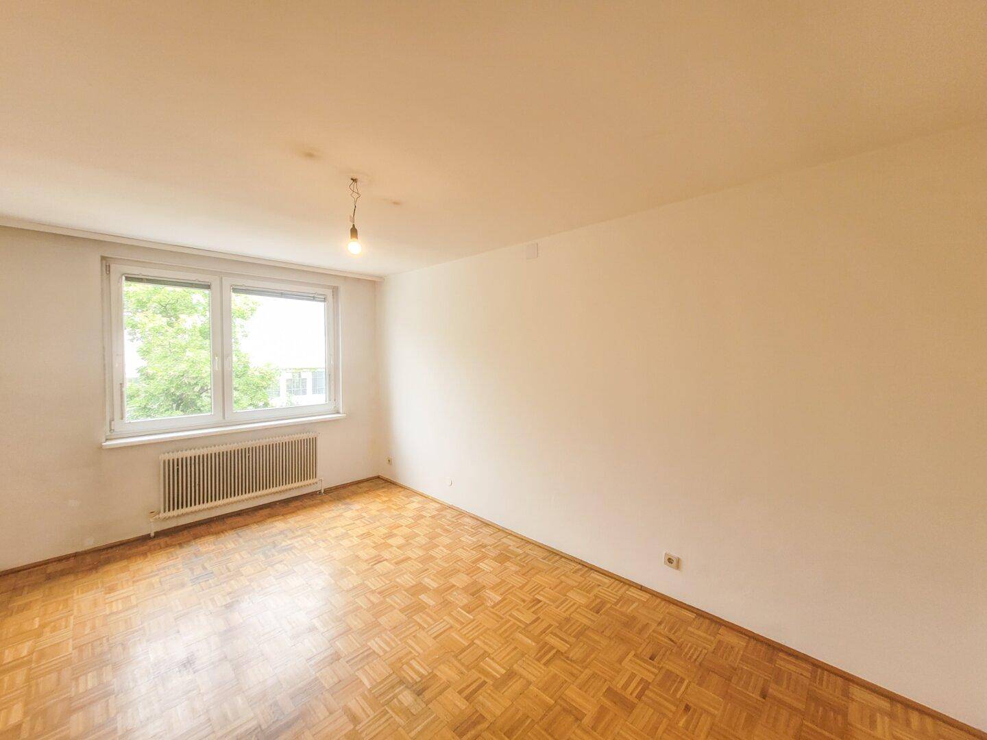 Geräumiges Zimmer mit Parkettboden, weißen Wänden und einem Fenster mit Blick auf Bäume.