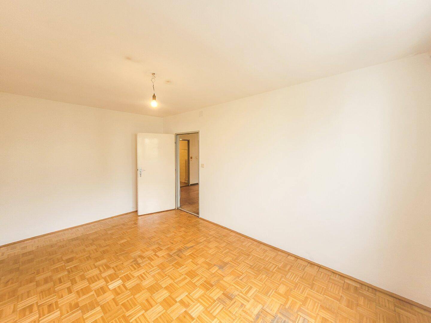 Leeres Zimmer mit Parkettboden und einer offenen Tür, die den Zugang zu anderen Räumen ermöglicht.