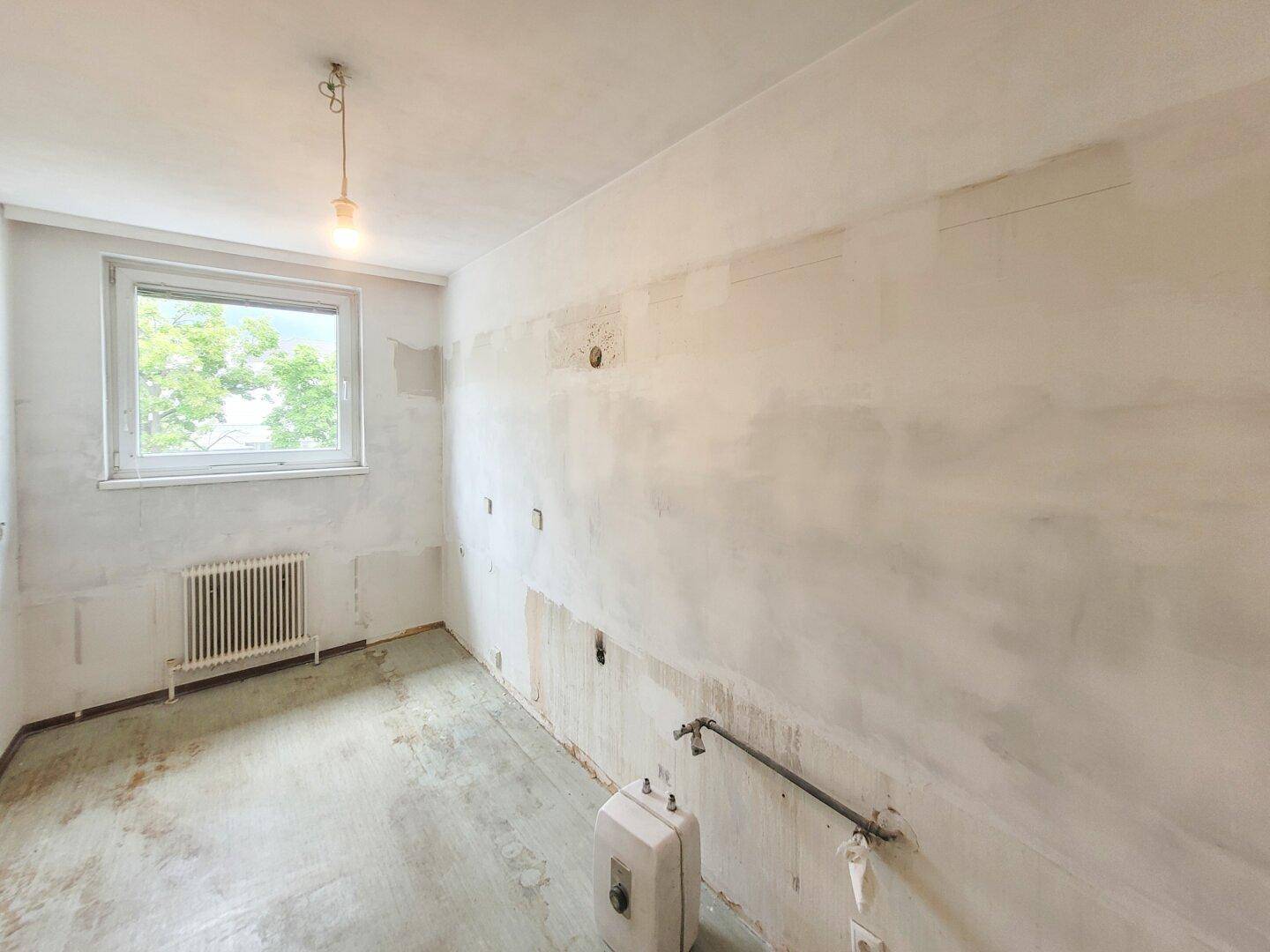 Renovierungsbedürftige Küche mit freigelegten Wänden, Betonboden und Fensterblick ins Grüne.