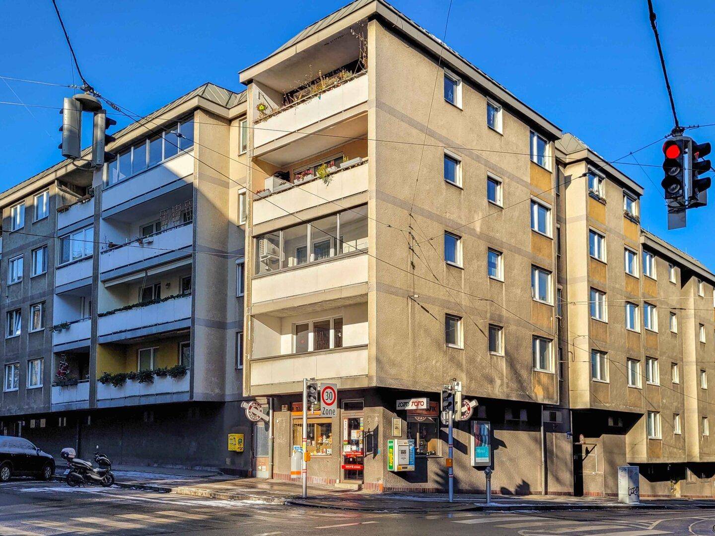Mehrstöckiges Wohngebäude an einer Straßenecke mit Balkonen und Geschäften im Erdgeschoss.