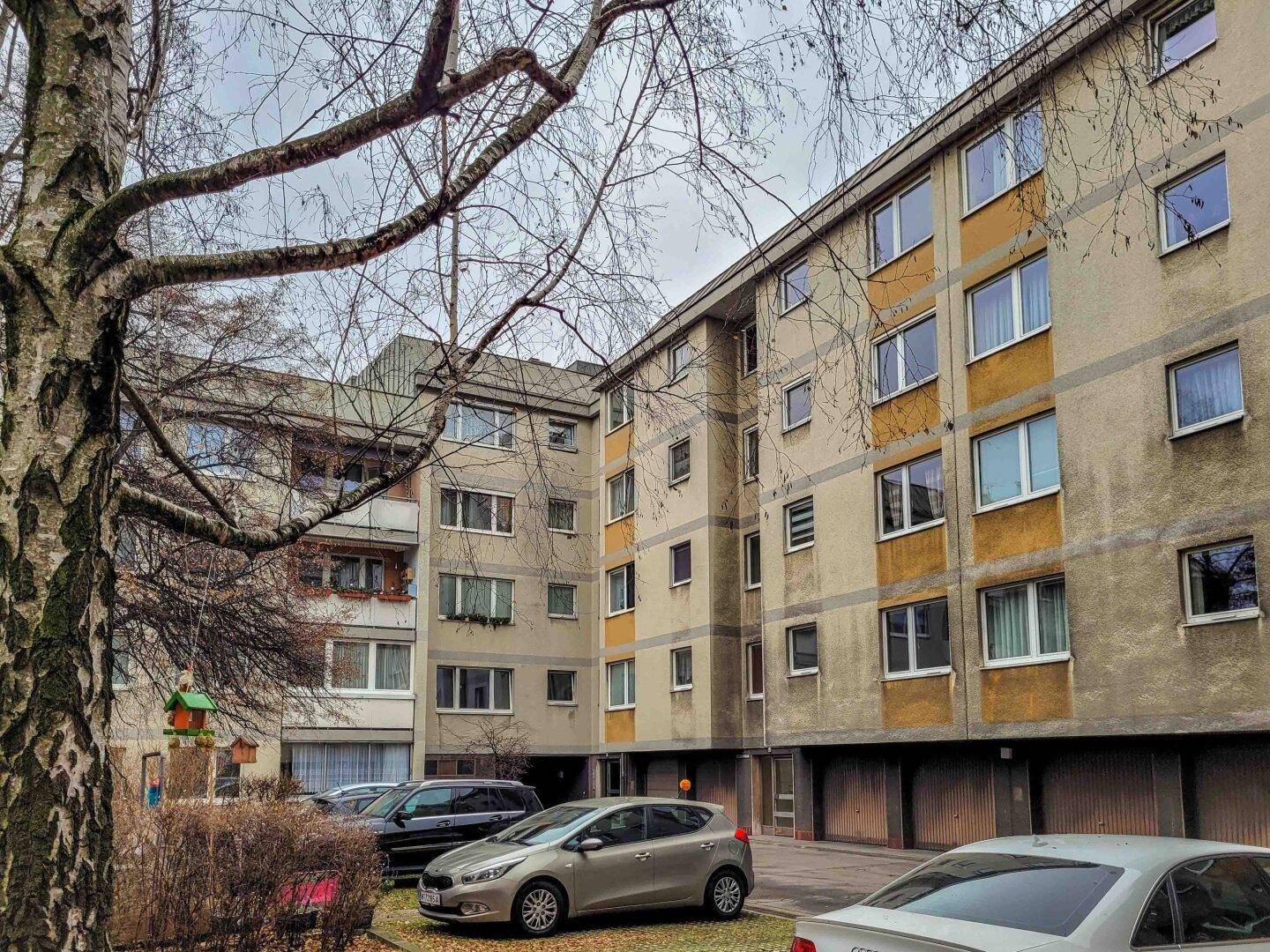 Mehrgeschossiges Wohnhaus mit Parkplätzen und Bäumen im begrünten Außenbereich.