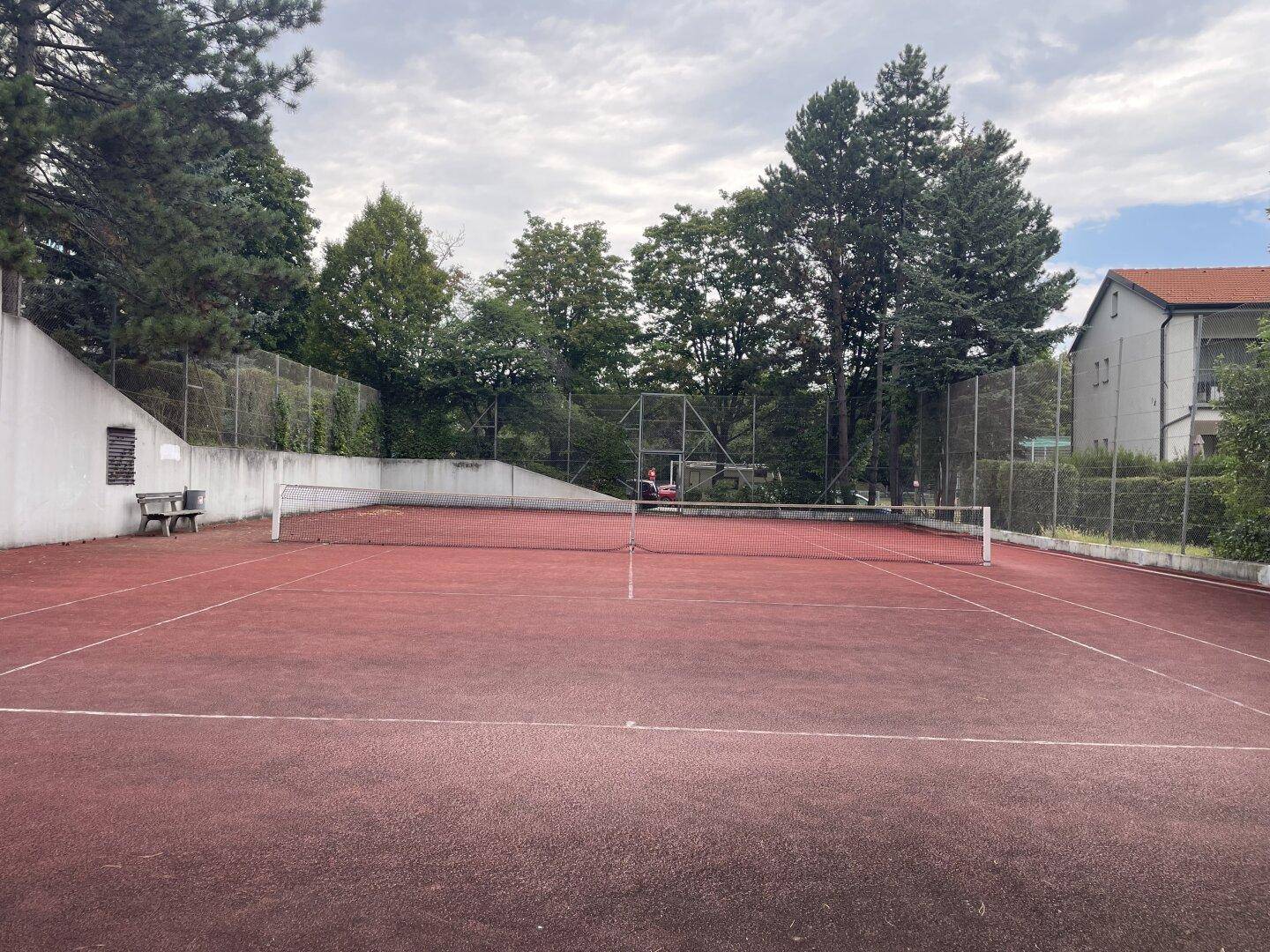 Roter Tennisplatz im Freien, umgeben von Bäumen und einem Zaun.
