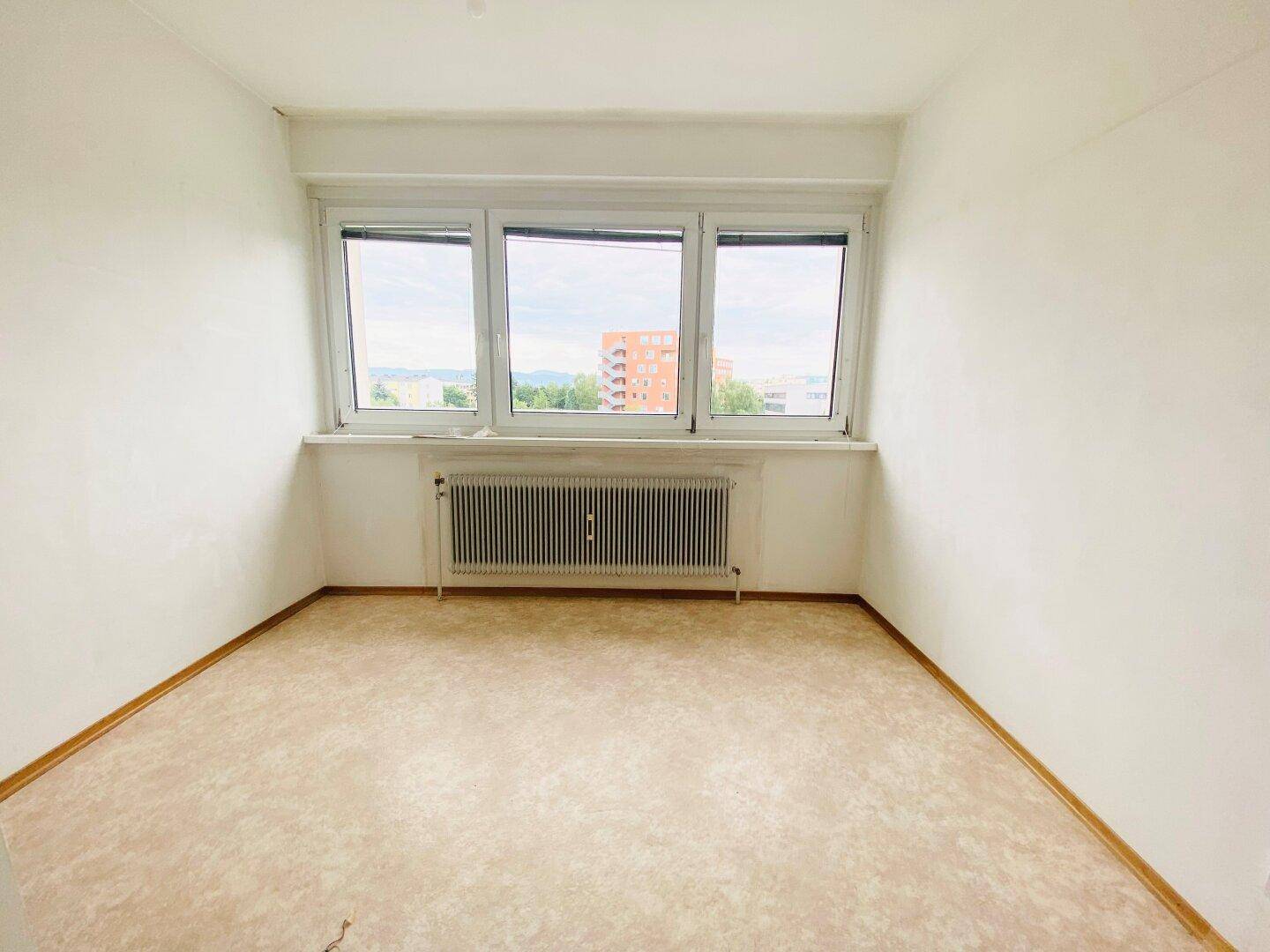 Helles Schlafzimmer mit hellem Boden, weißen Wänden und einem großen Fenster mit Blick ins Grüne.