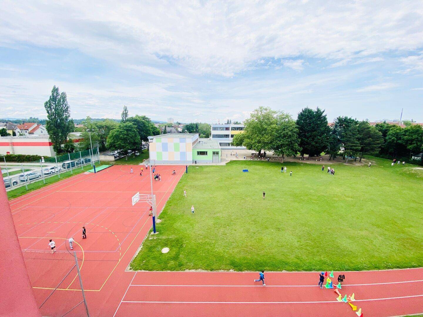 Weitläufige Sportanlagen mit Laufbahn, Basketballplatz und großer Rasenfläche, umgeben von Bäumen.