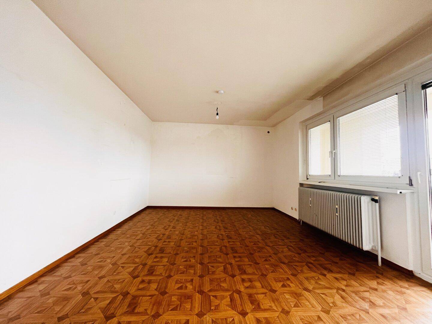 Geräumiges Wohnzimmer mit Fischgrätparkettboden, weißen Wänden und Zugang zu einem Balkon.