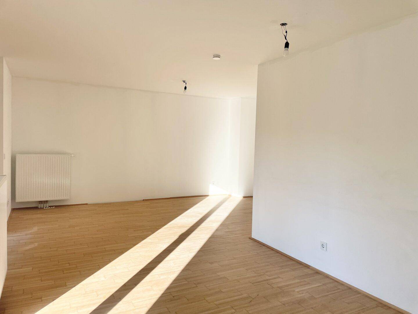 Lichtdurchflutetes Wohnzimmer mit hellem Parkettboden und schlichten weißen Wänden.