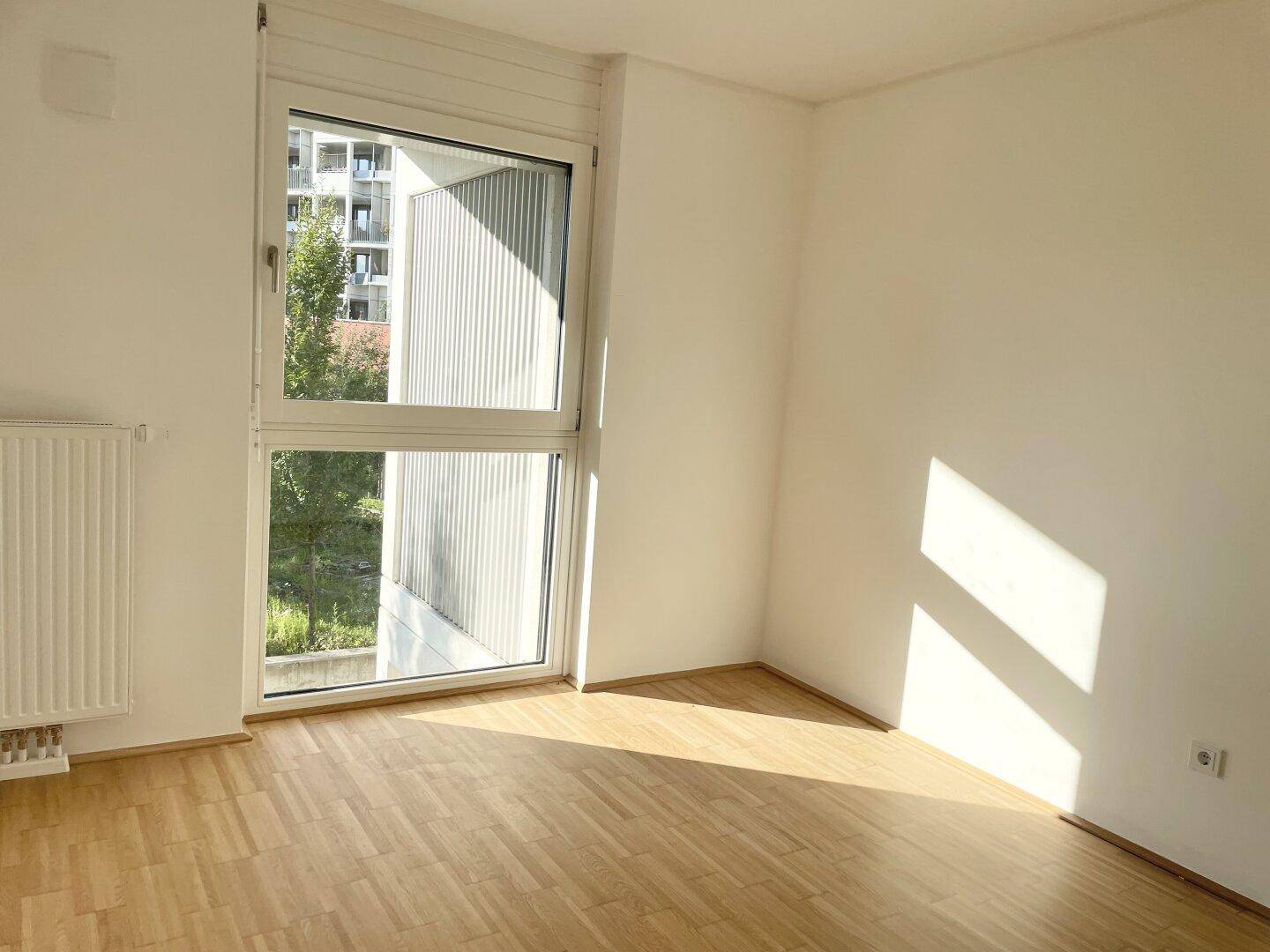 Helles Zimmer mit Parkettboden, Heizkörper und großem Fenster mit Blick ins Grüne.