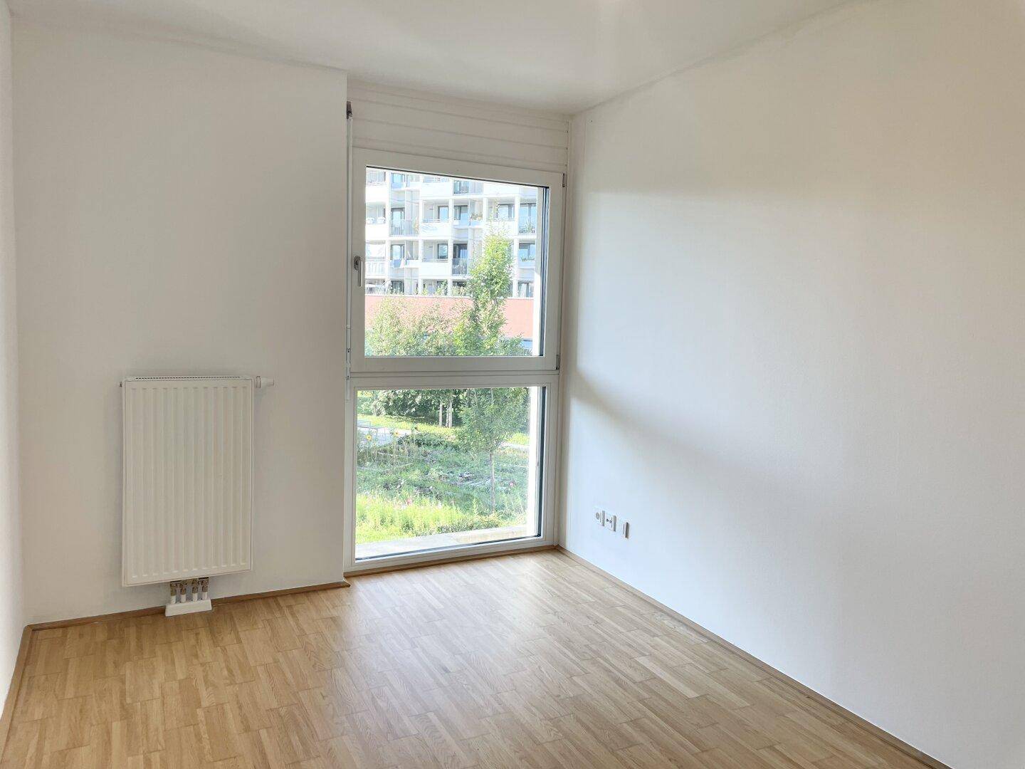 Freundliches Zimmer mit Parkettboden, Heizkörper und Fenster mit Blick auf die Umgebung.