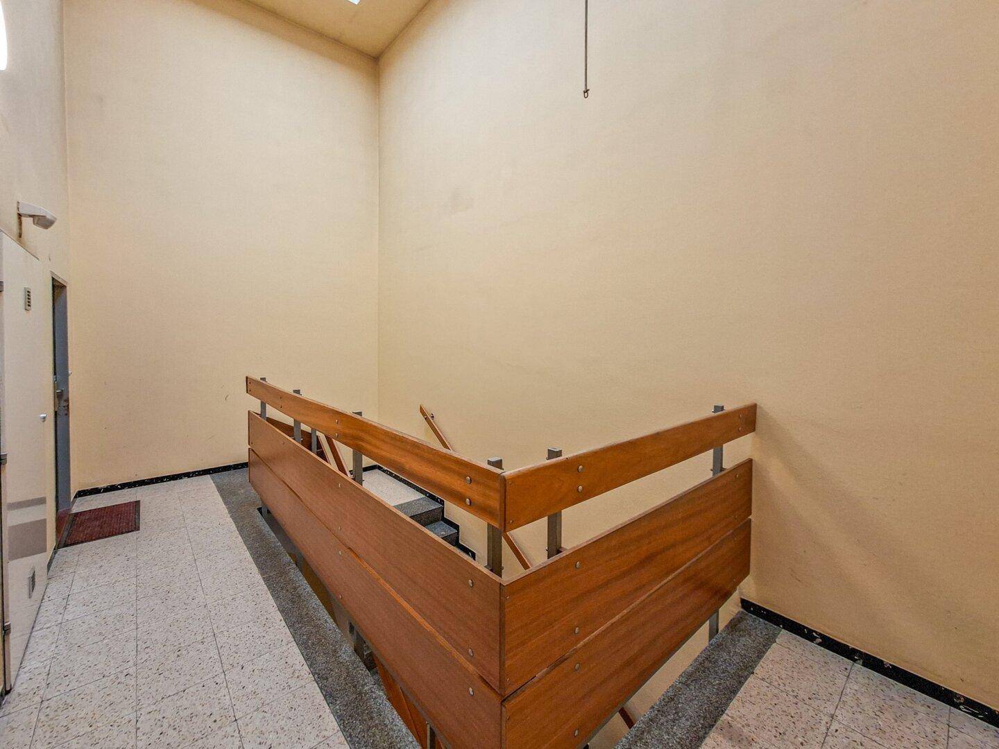 Treppenhaus mit Holzgeländer und Terrazzoboden, das zu den oberen Etagen führt.