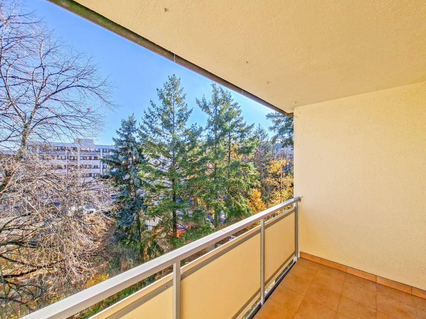 Balkon mit Blick auf grüne Bäume und umliegende Gebäude unter blauem Himmel.
