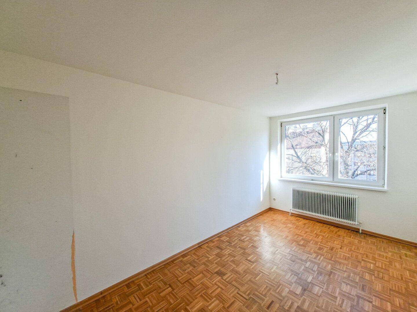 Leeres Zimmer mit Fischgrätparkett, weißen Wänden und großem Fenster für viel Tageslicht.
