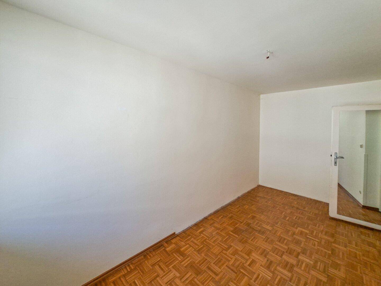 Leeres Zimmer mit Fischgrätparkett und weißen Wänden, das auf eine Renovierung wartet.
