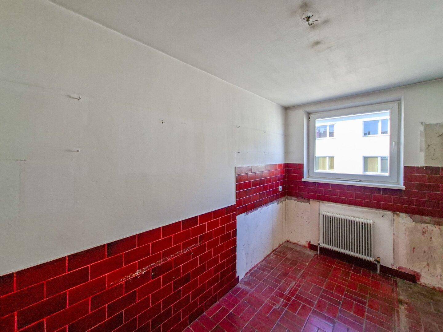 Küche in Abrisszustand mit roten Fliesen an Wand und Boden sowie einem Fenster.
