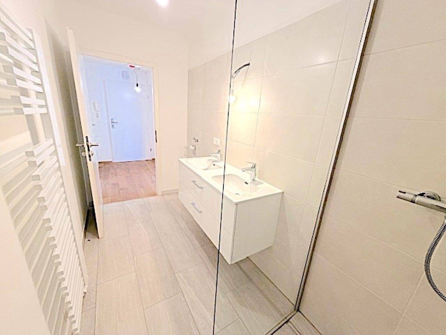 Modernes Badezimmer mit hellen Fliesen, Doppelwaschbecken, begehbarer Dusche und Handtuchheizkörper.