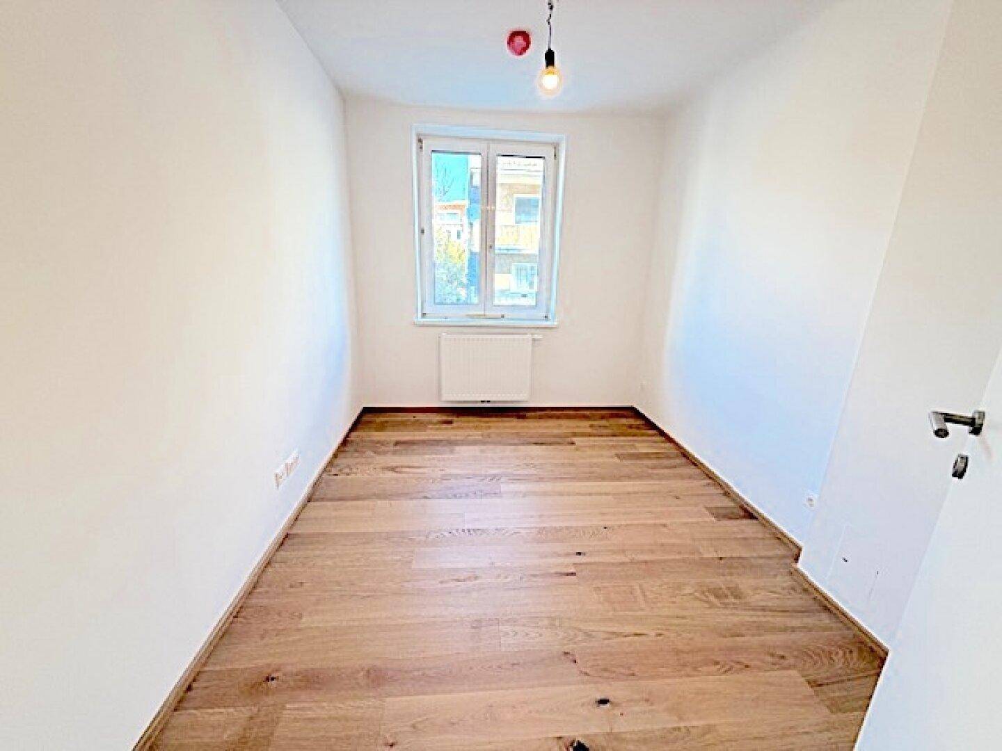 Leeres Zimmer mit hellem Holzparkettboden und einem Fenster für Tageslicht.