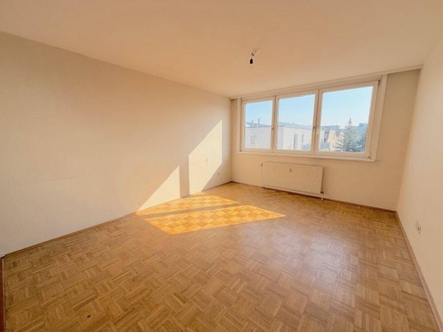 Sonniges, leeres Zimmer mit Parkettboden und breiten Fenstern, die einen hellen Raum schaffen.