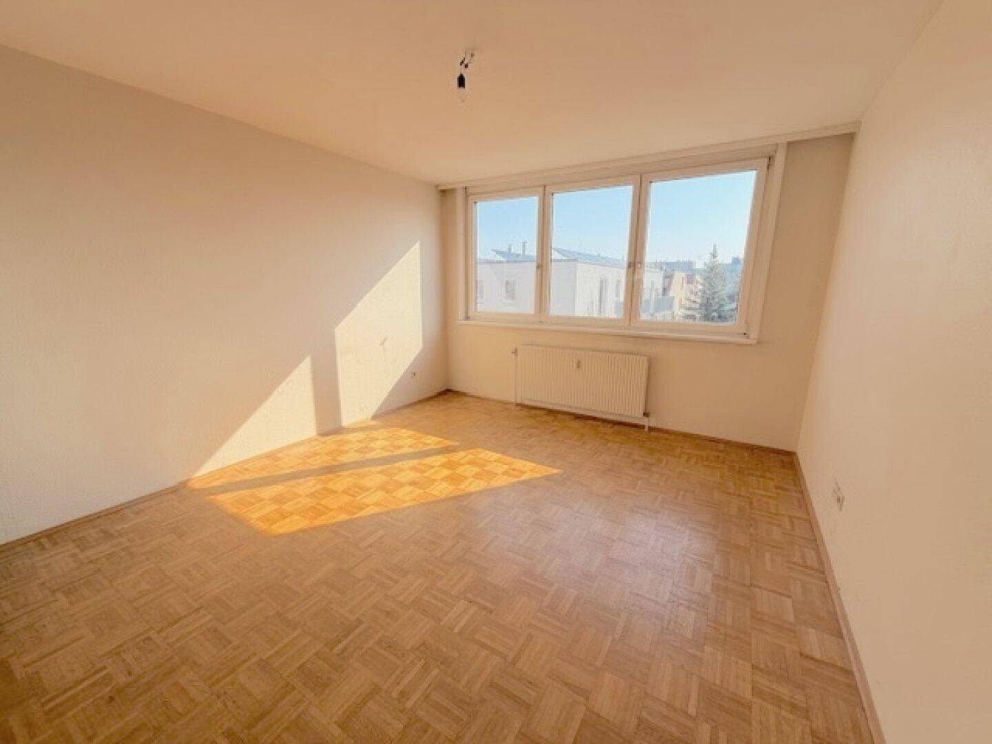 Großes, leeres Zimmer mit Parkettboden und hellen Fenstern, die viel Tageslicht hereinlassen.