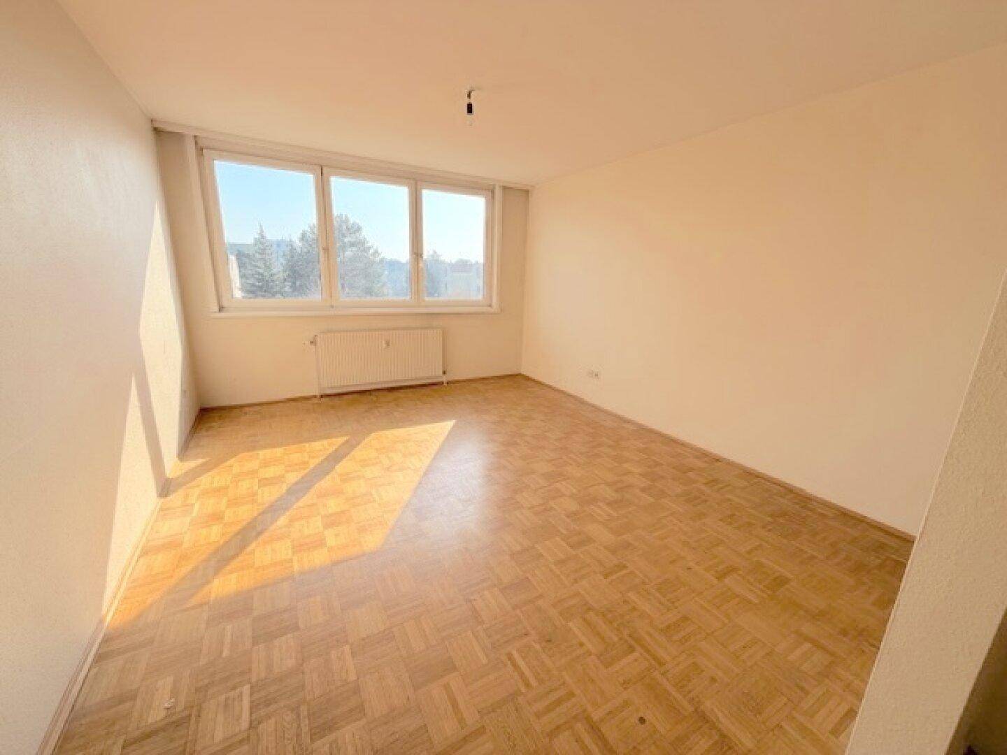 Geräumiges, leeres Zimmer mit hellem Parkettboden und großen Fenstern, durch die Sonnenlicht fällt.