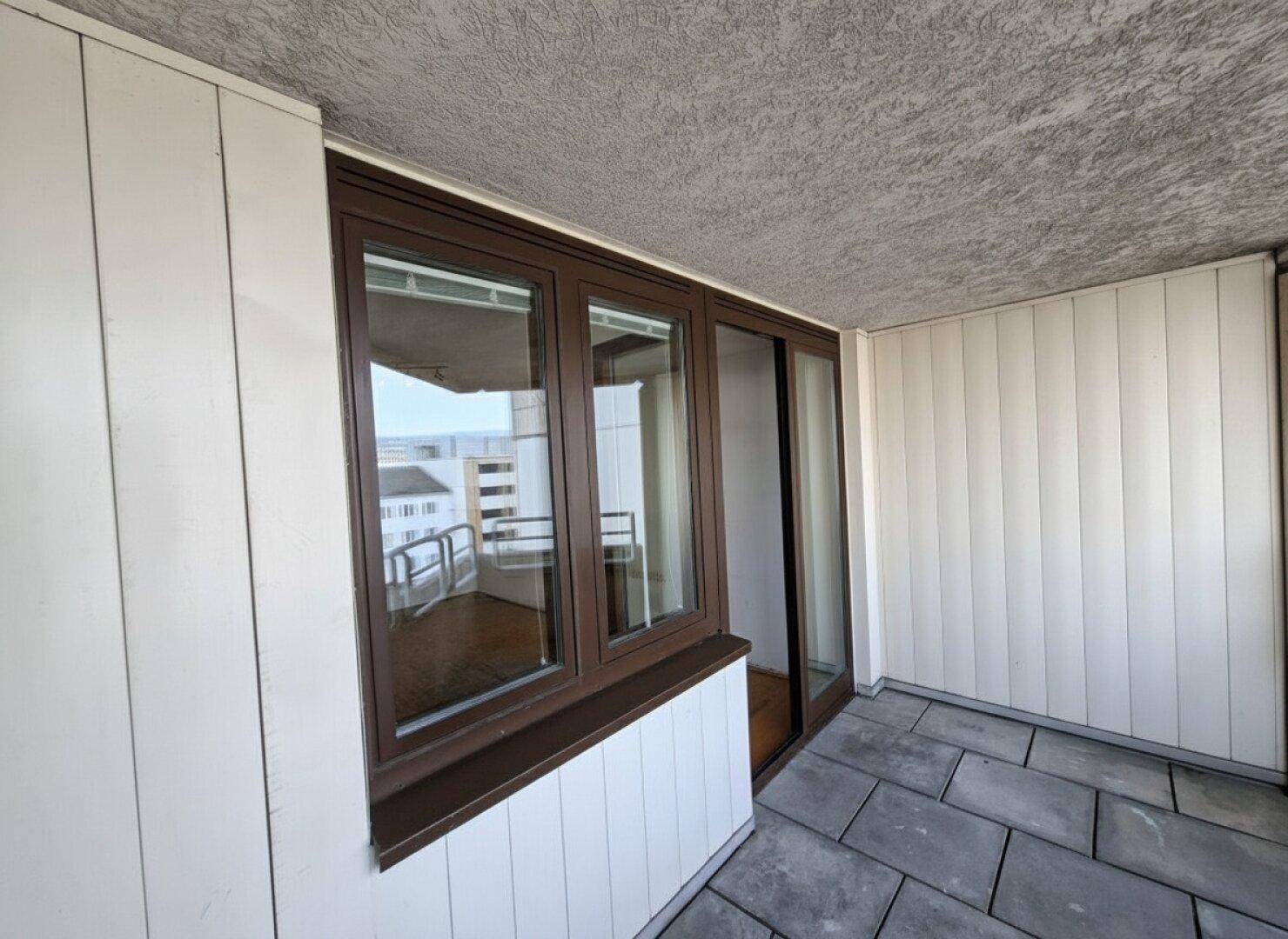 Balkon mit Fliesenboden und braunen Fensterrahmen, bietet einen Blick auf die Umgebung.