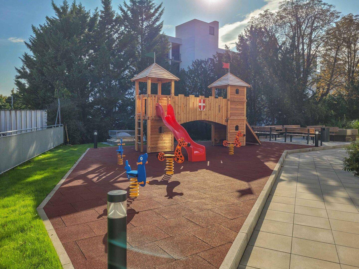 Kinderspielplatz mit Rutsche und Schaukeln, umgeben von Grünflächen und Bäumen.